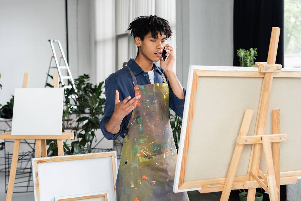 African american artist in dirty apron talking on smartphone near canvas in studio  - Fotografie, Obrázek