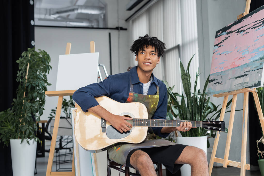 Kirli önlüklü gülümseyen Afrikalı Amerikalı sanatçı stüdyoda resim sehpası yakınlarında akustik gitar çalıyor.  - Fotoğraf, Görsel