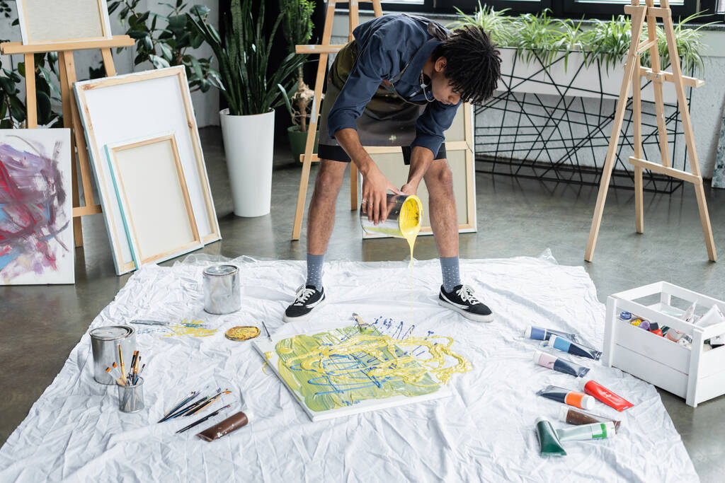 Νεαρός αφροαμερικανός καλλιτέχνης ρίχνει χρώμα σε καμβά και ύφασμα σε εργαστήριο τέχνης  - Φωτογραφία, εικόνα