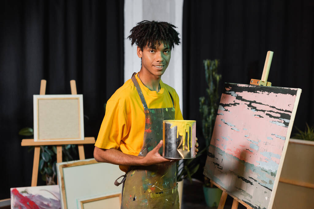 Smiling african american artist holding jar with paint near drawing in workshop  - Photo, image