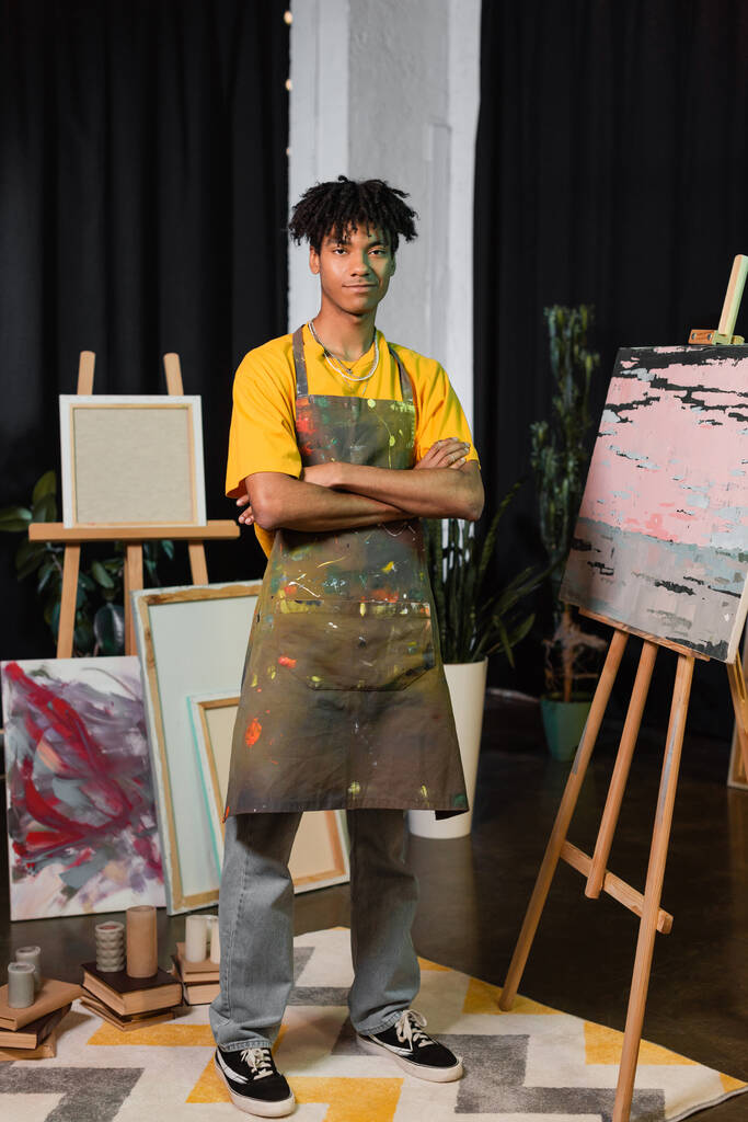 Smiling african american artist crossing arms near paintings in studio  - Foto, imagen