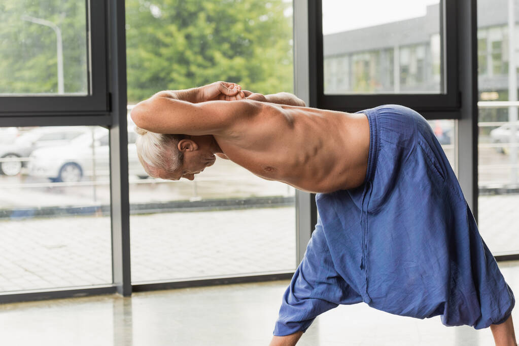 flexible and grey haired man doing standing forward bend pose in yoga studio  - Foto, Imagen