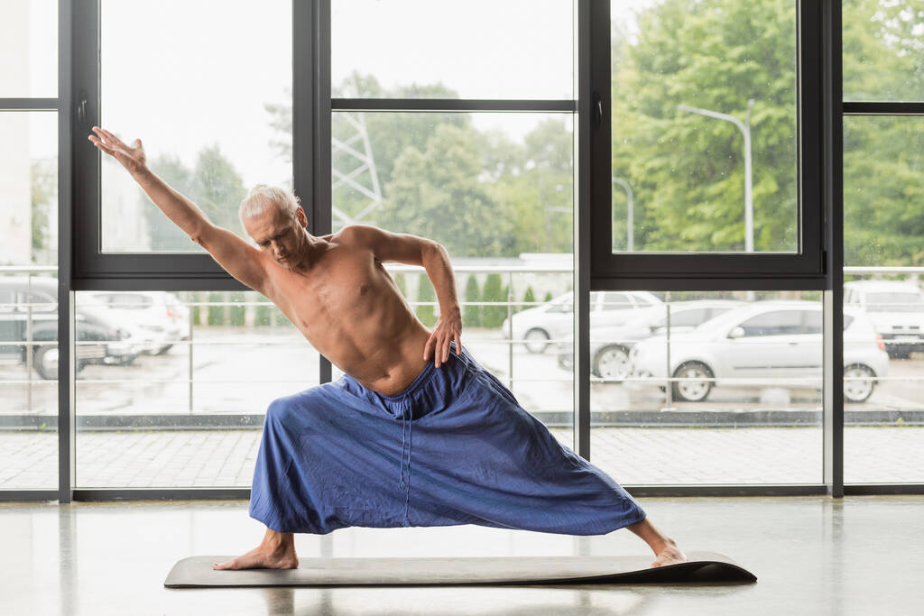 hombre sin camisa y de pelo gris haciendo postura de ángulo lateral extendido en estudio de yoga  - Foto, imagen