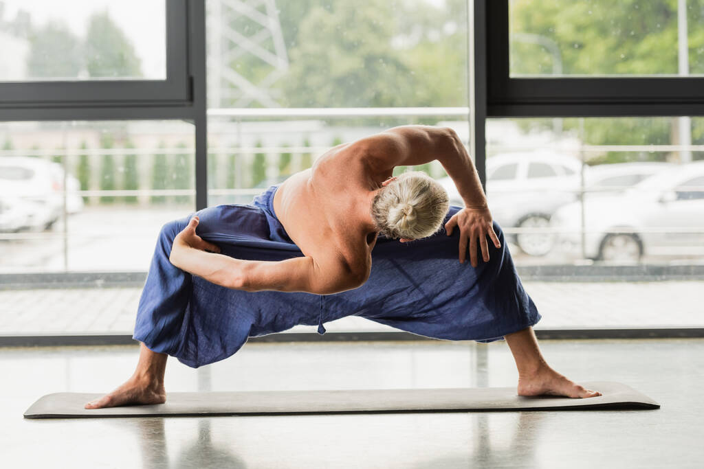 седовласый мужчина делает широкий поворот вперед ногами, практикуя йогу  - Фото, изображение