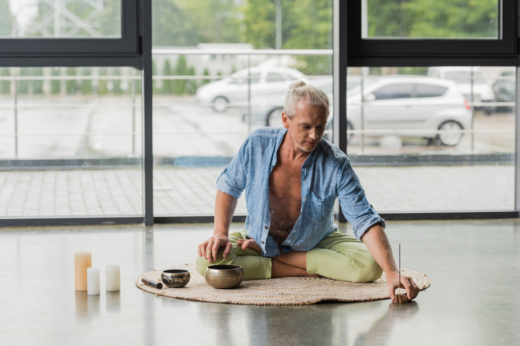 grey haired man moving incense stick near Tibetan singing bowls and candles in yoga studio  - Photo, image