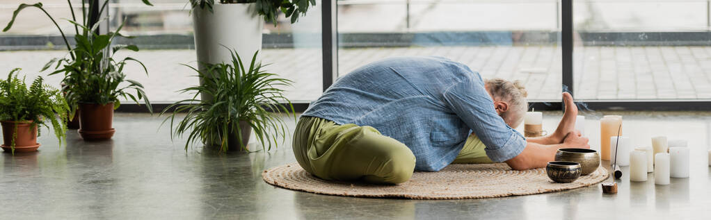 grey haired man stretching back near Tibetan singing bowls and candles in yoga studio, banner  - Foto, Imagem
