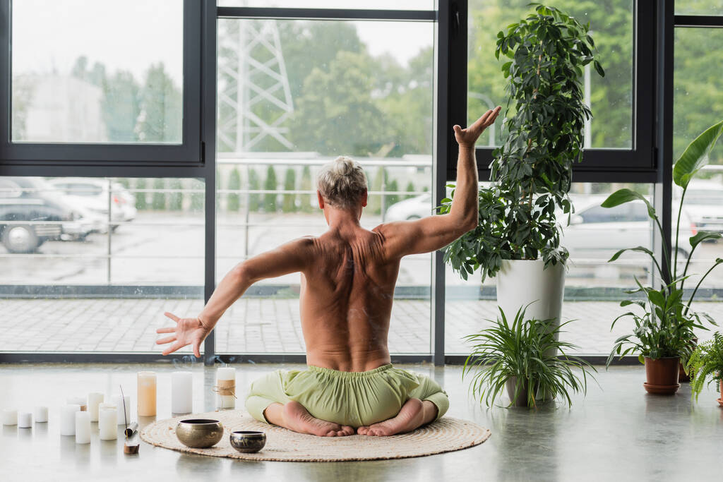 back view of shirtless man gesturing while sitting in thunderbolt yoga pose near candles and Tibetan singing bowls  - Фото, зображення