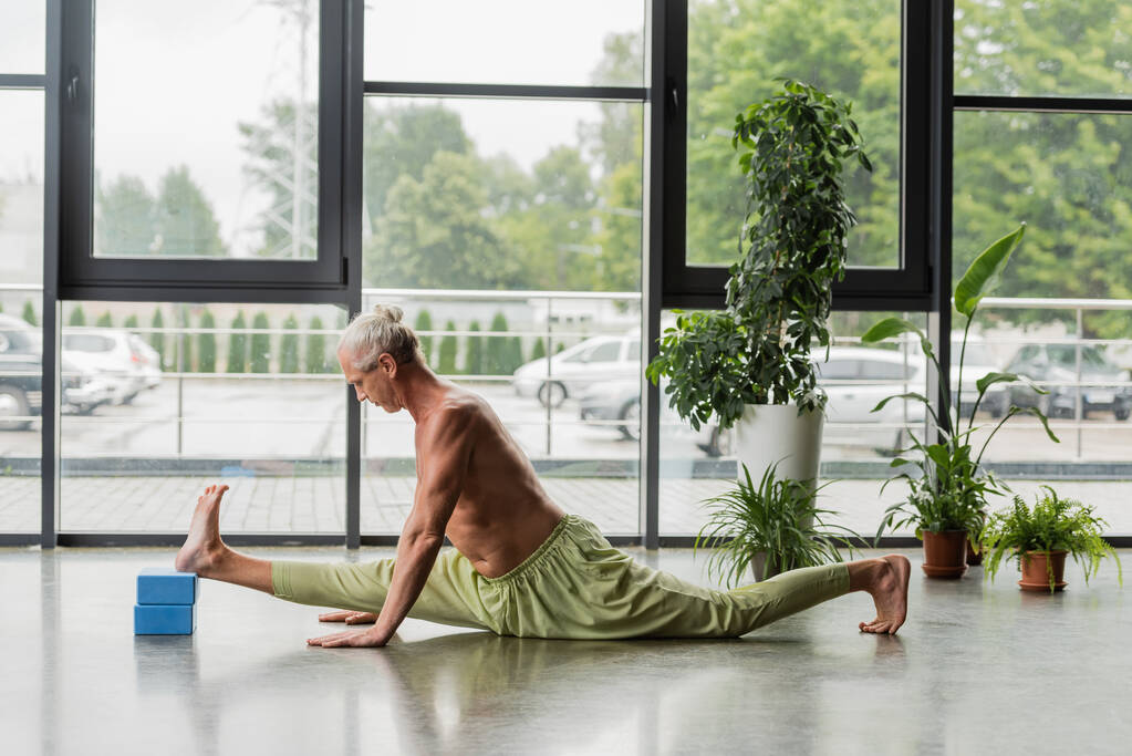 zijaanzicht van shirtloze en grijsharige man doet split op blauwe yoga schuim blokken in studio  - Foto, afbeelding