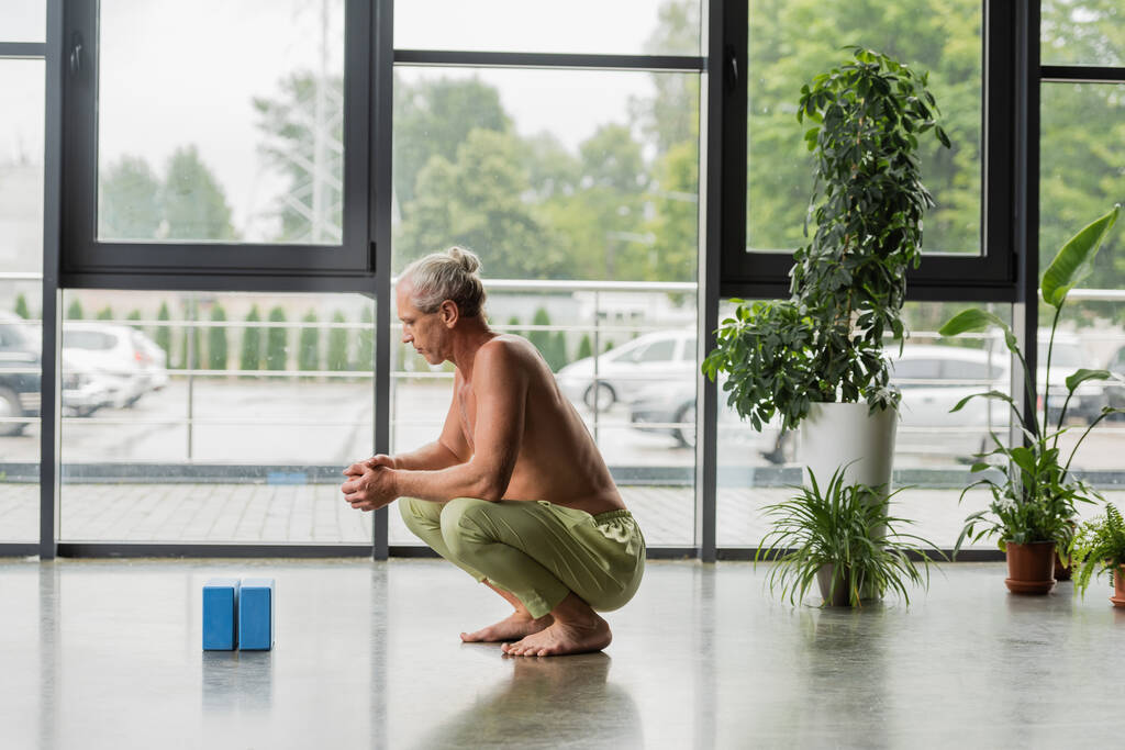 vue latérale de l'homme sans chemise et aux cheveux gris assis avec les mains serrées et regardant des blocs de mousse de yoga bleu en studio  - Photo, image