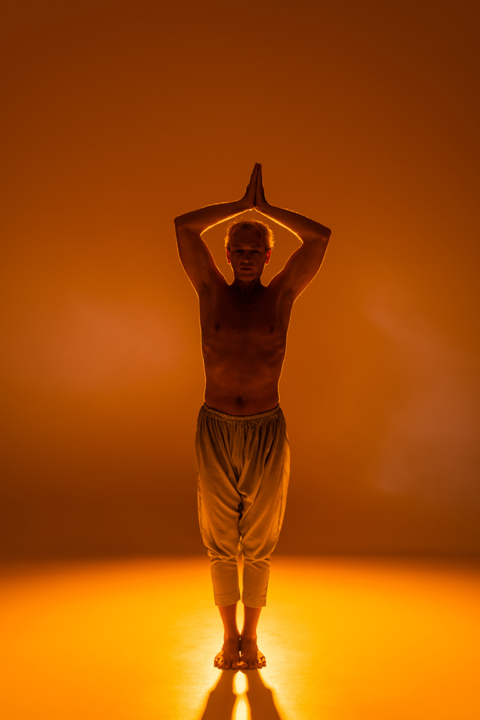 full length of shirtless man in pants standing in warrior pose on orange background  - Foto, imagen