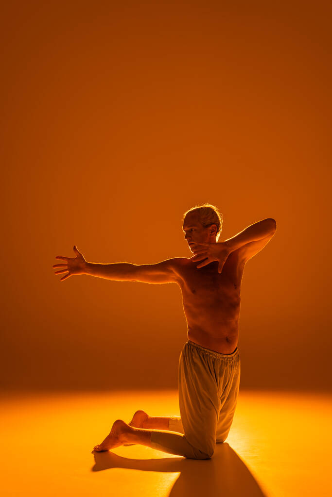 full length of shirtless man standing on knees while meditating on brown background  - Foto, Imagen