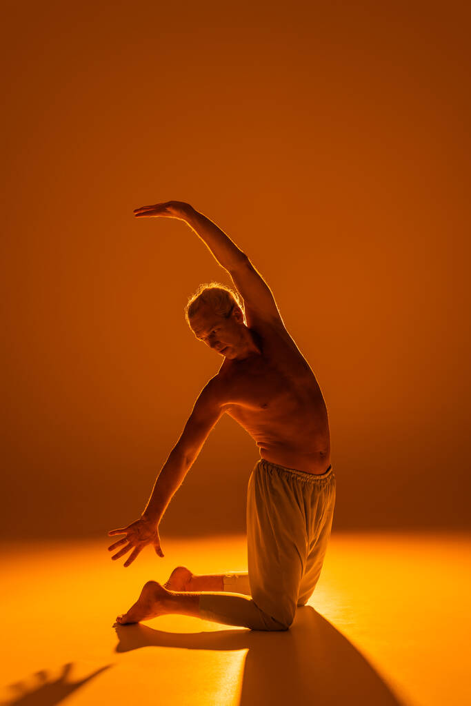 full length of shirtless man standing on knees while practicing yoga on brown background  - Foto, Bild