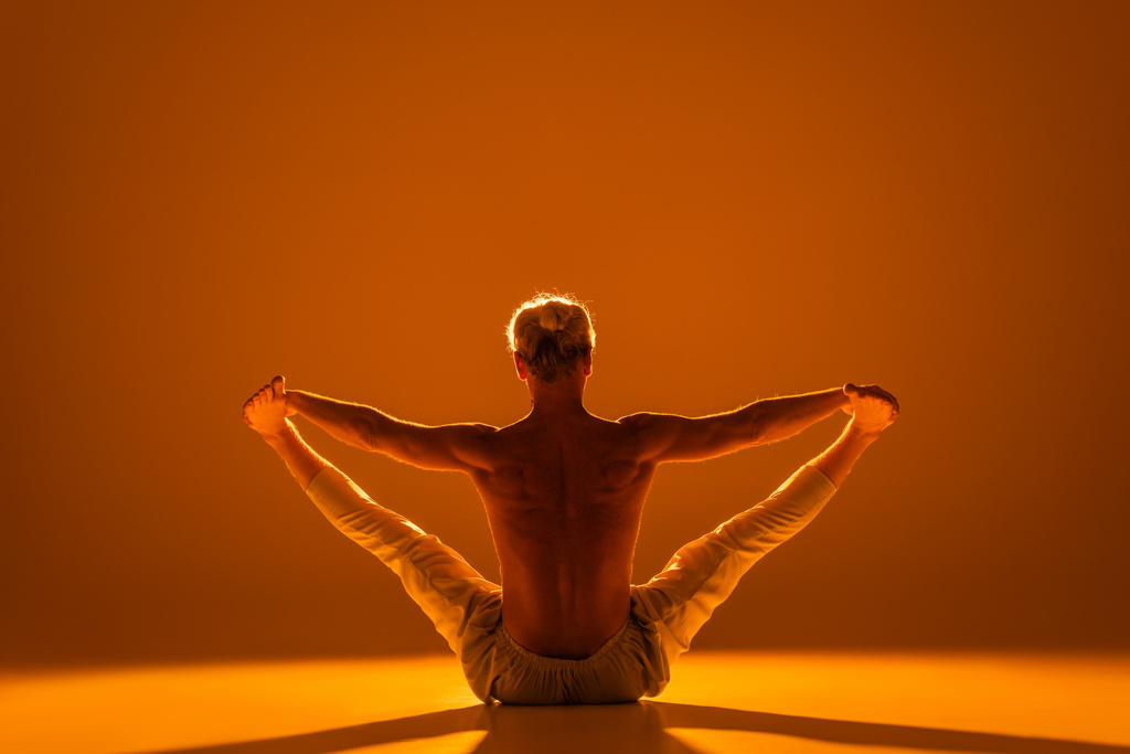 achteraanzicht van shirtless man doen zittende handen naar tenen yoga pose op bruin  - Foto, afbeelding