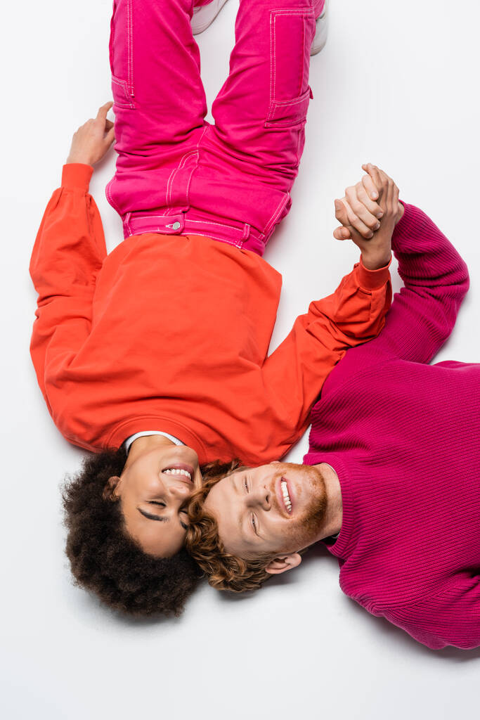 top view of pleased redhead man and curly african american woman in magenta color clothes lying on white  - Zdjęcie, obraz