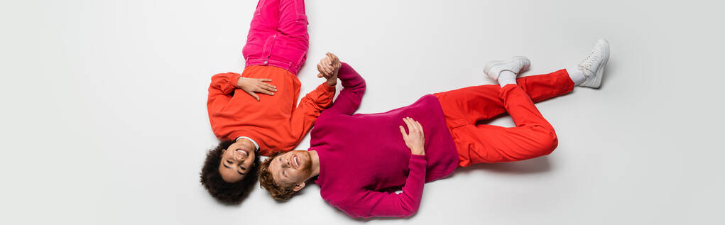 top view of happy redhead man and joyful african american woman in magenta color clothes lying on white, banner  - Photo, Image
