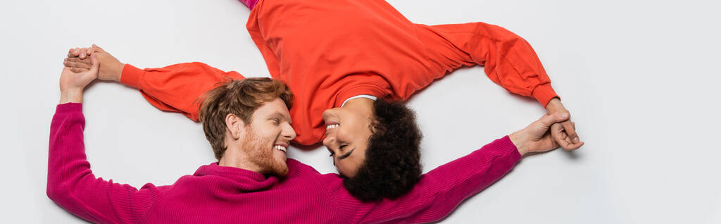 top view of happy redhead man in magenta color clothes and curly african american woman holding hands on white, banner  - Photo, Image