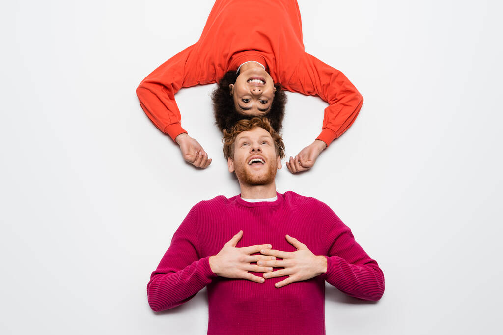 top view of happy redhead man in magenta color clothes and excited african american woman lying on white  - Foto, immagini