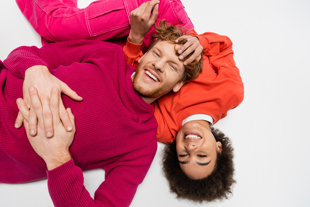 вид сверху на позитивную африканскую американку лежащую с рыжеволосым мужчиной в пурпурной цветовой одежде на белом  - Фото, изображение