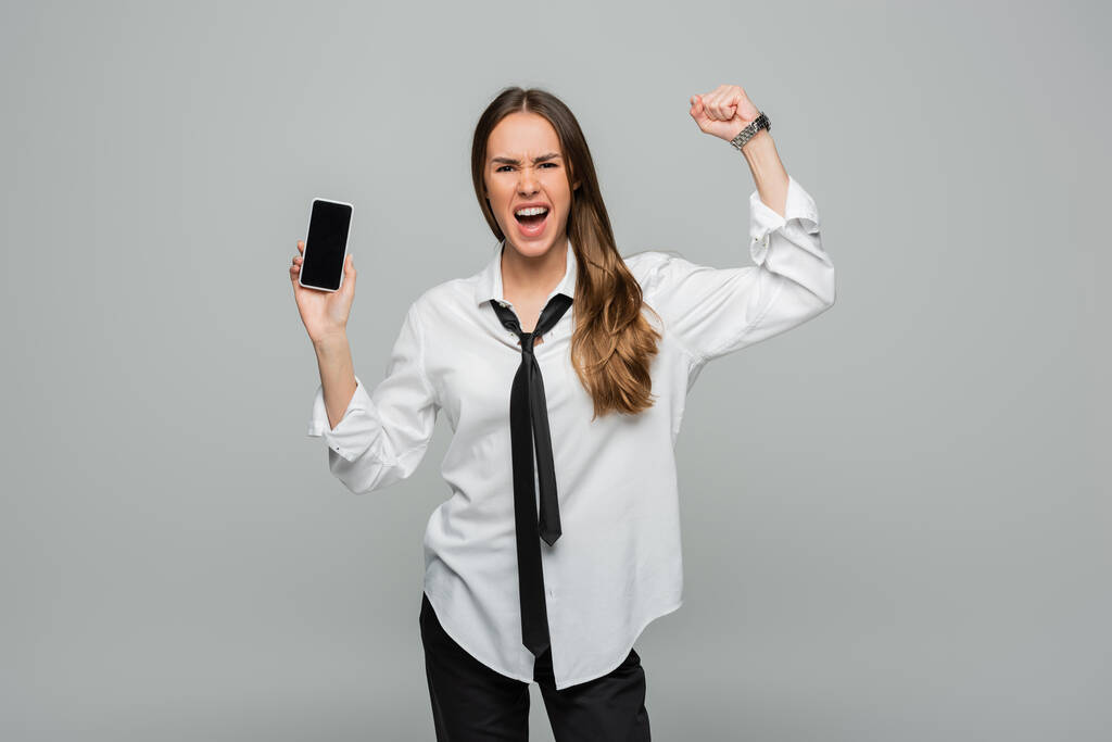 femme émotionnelle en chemise et cravate debout avec le poing serré et tenant smartphone avec écran blanc isolé sur gris, égalité des sexes - Photo, image