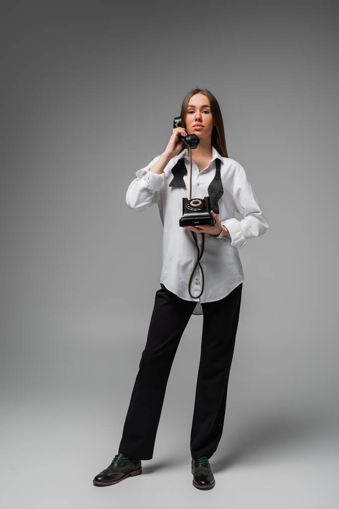 full length of young woman in white shirt with tie and pants using retro telephone on grey  - 写真・画像