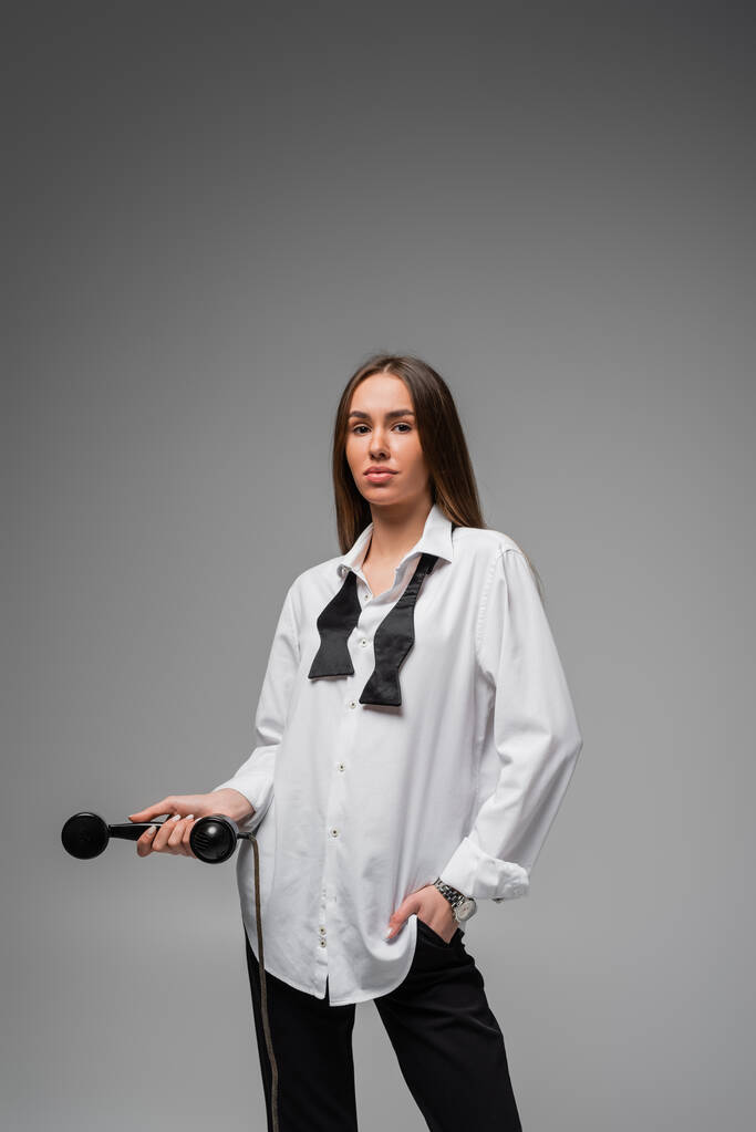 молодая женщина в белой рубашке с галстуком и брюками держа ретро телефон, позируя на сером  - Фото, изображение