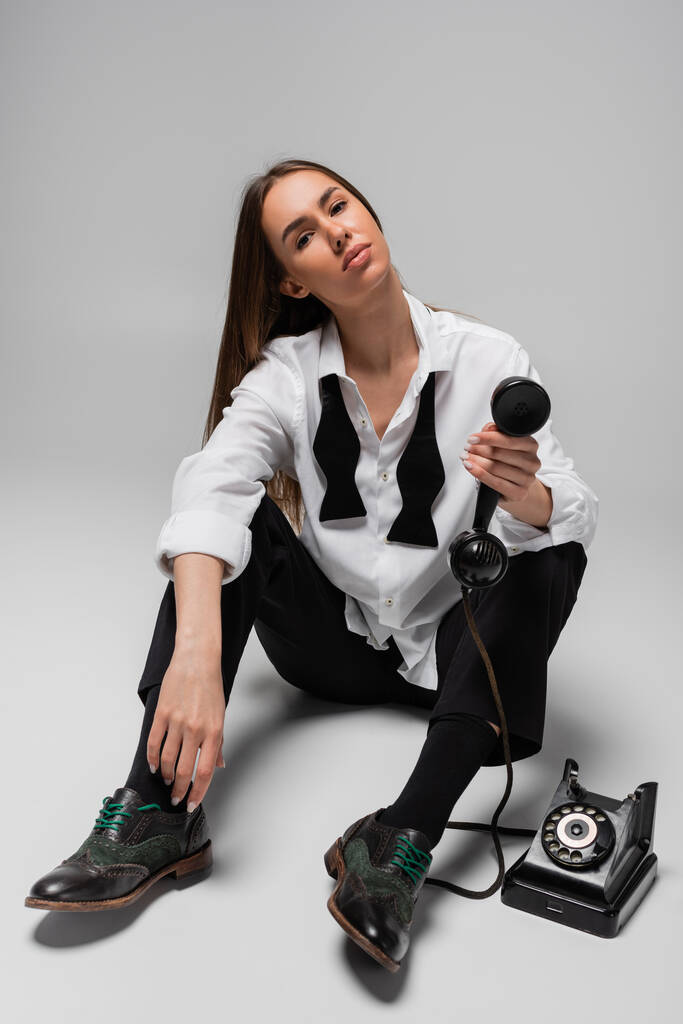 young woman in shirt with tie and pants holding retro telephone while sitting on grey  - Photo, Image