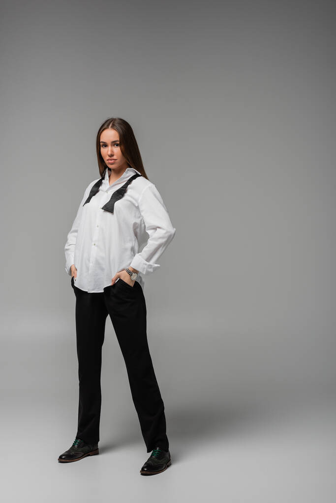 longitud completa de mujer confiada en camisa con corbata y pantalones negros de pie con las manos en los bolsillos en gris, concepto de igualdad de género  - Foto, Imagen