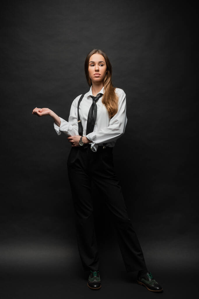 full length of brunette woman in formal wear with suspenders standing and looking away on black  - Foto, afbeelding