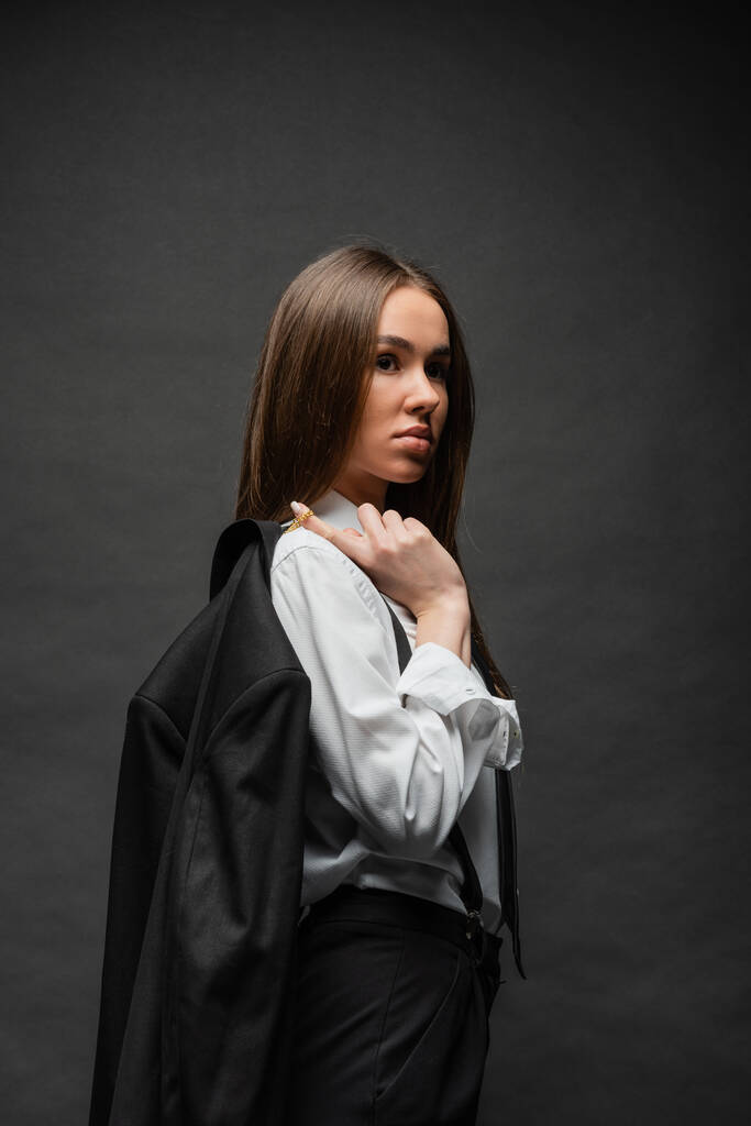 brunette woman with long hair standing in formal wear and holding blazer on black  - Foto, Imagem