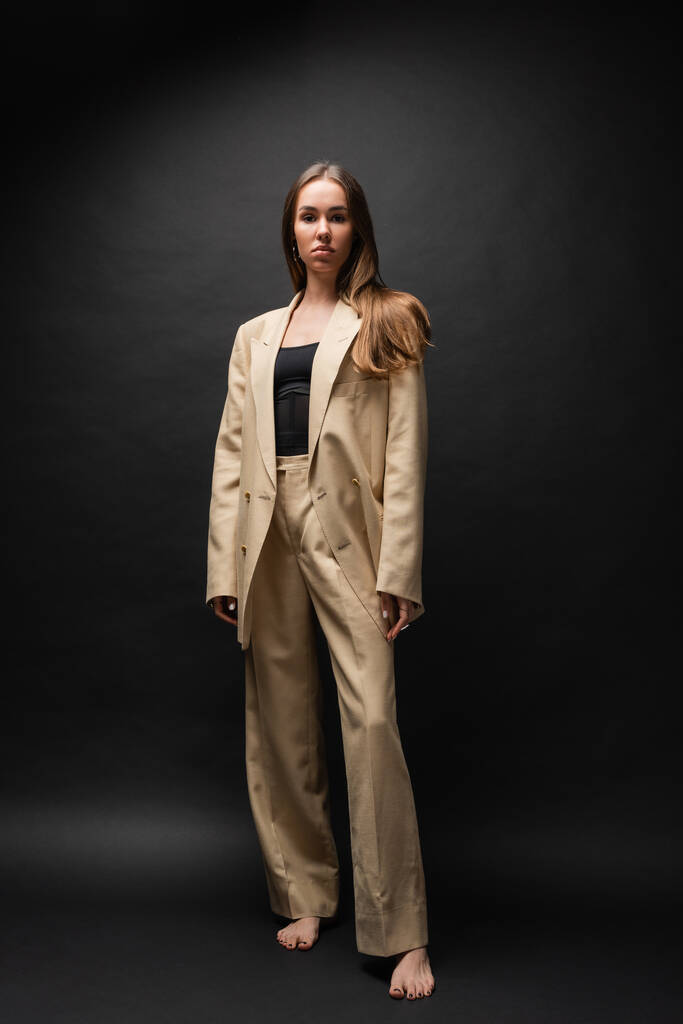 full length of barefoot woman in beige suit standing and looking at camera on black  - Fotoğraf, Görsel
