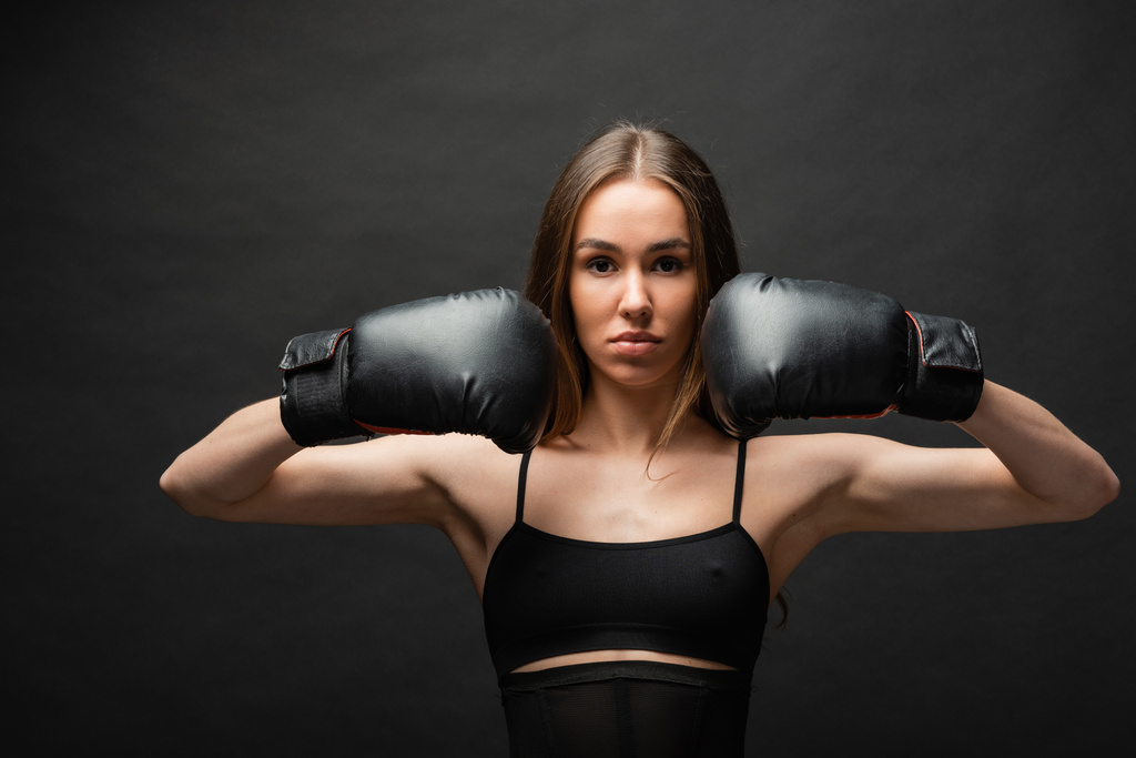 brunette and strong young woman in top posing in boxing gloves on black background  - 写真・画像