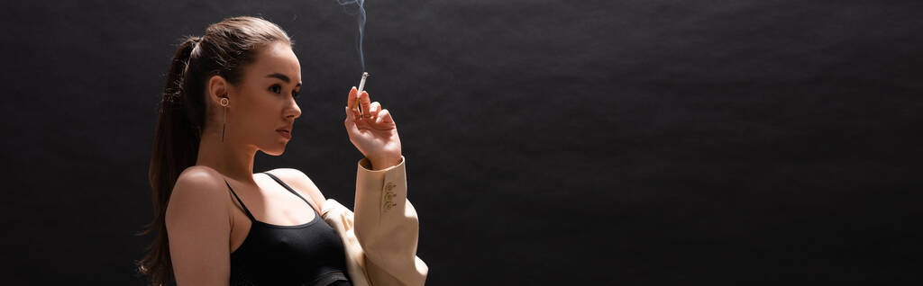 young brunette woman in beige blazer holding cigarette while looking away on black background, banner  - Foto, afbeelding
