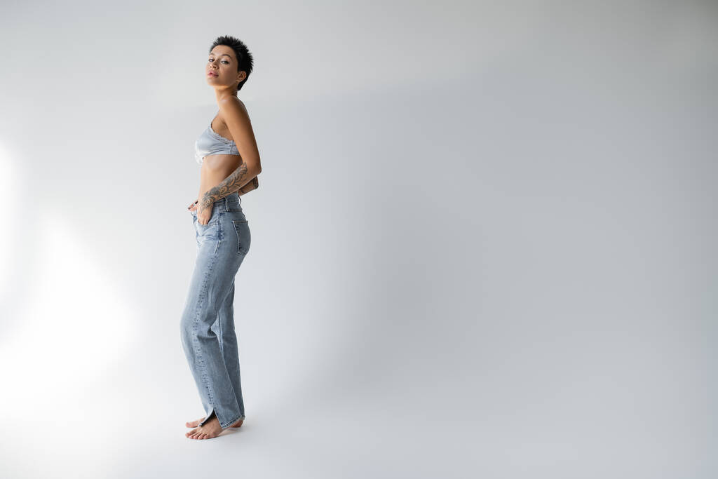 повна довжина босоніж брюнетка жінка в бюстгальтері і джинсах стоїть з рукою в кишені на сірому фоні
 - Фото, зображення