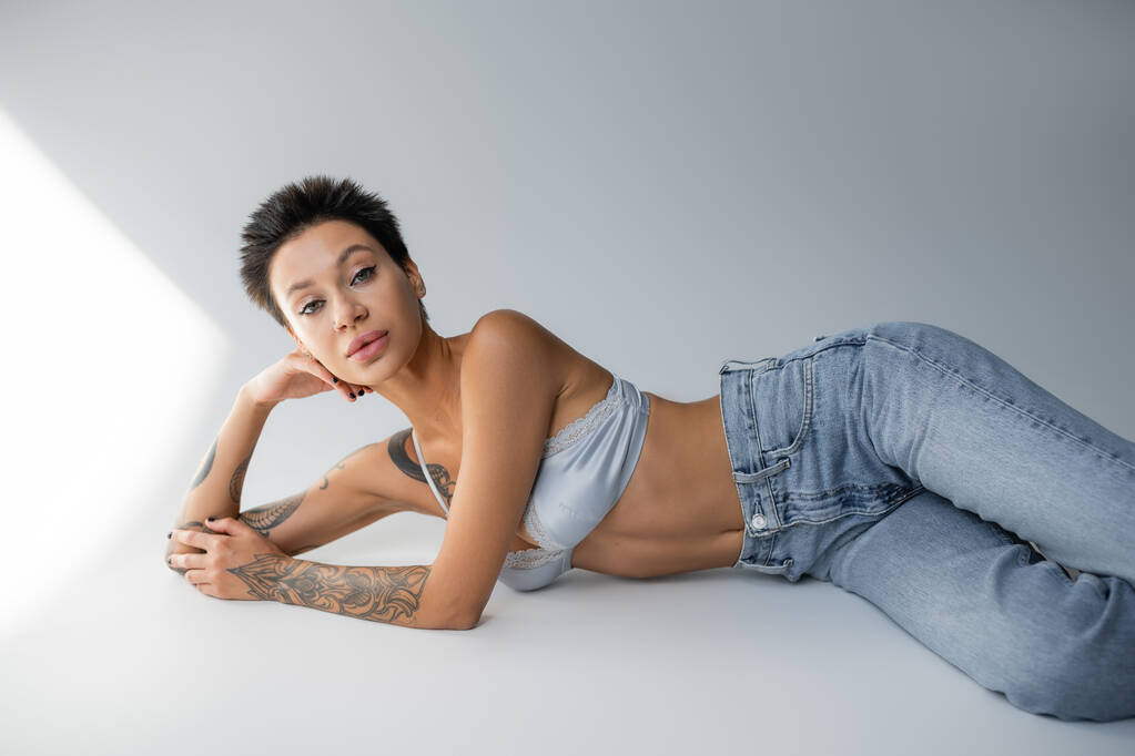short haired and tattooed woman in blue jeans with bra lying on grey background - Foto, Imagem