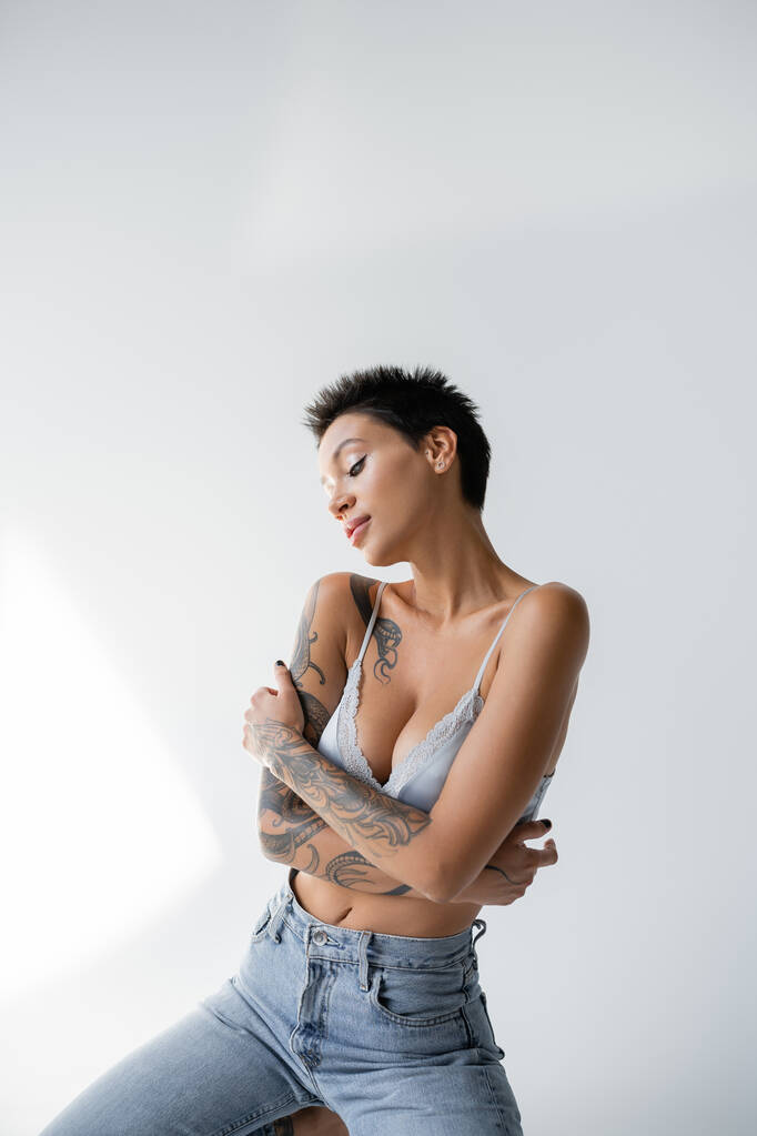 femme tatouée sensuelle en jeans et soutien-gorge en satin assis avec les bras croisés sur fond gris - Photo, image