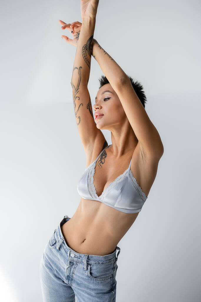 tattooed and slender woman in silk bra and jeans standing with raised hands on grey background - Photo, Image