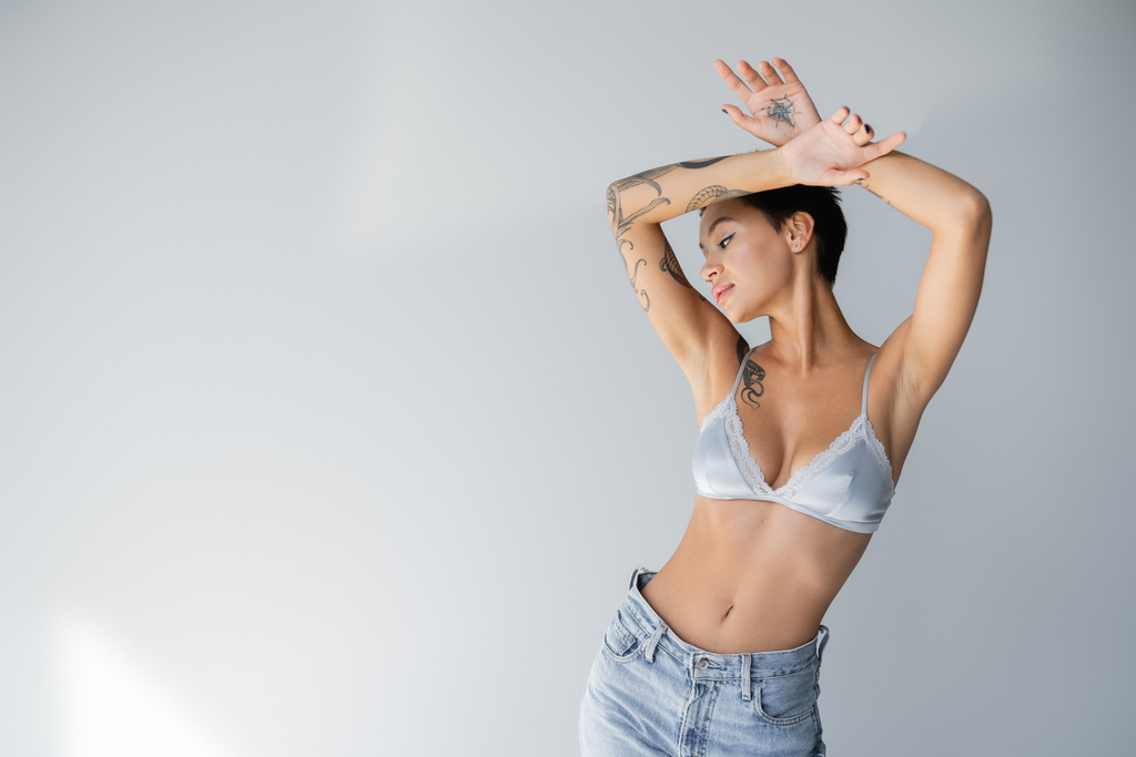 young and slender woman in satin bra and jeans posing with tattooed hands above head on grey background - Fotó, kép