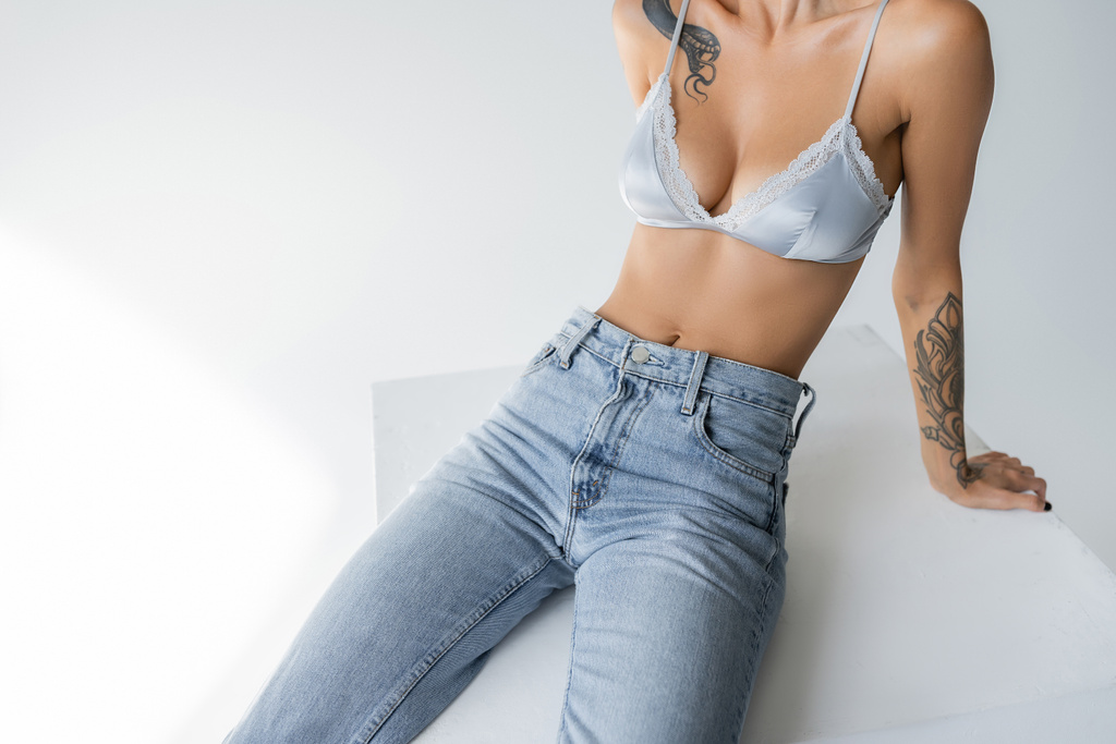 partial view of slender woman in blue jeans and silk bralette sitting on cube on grey background - Photo, Image