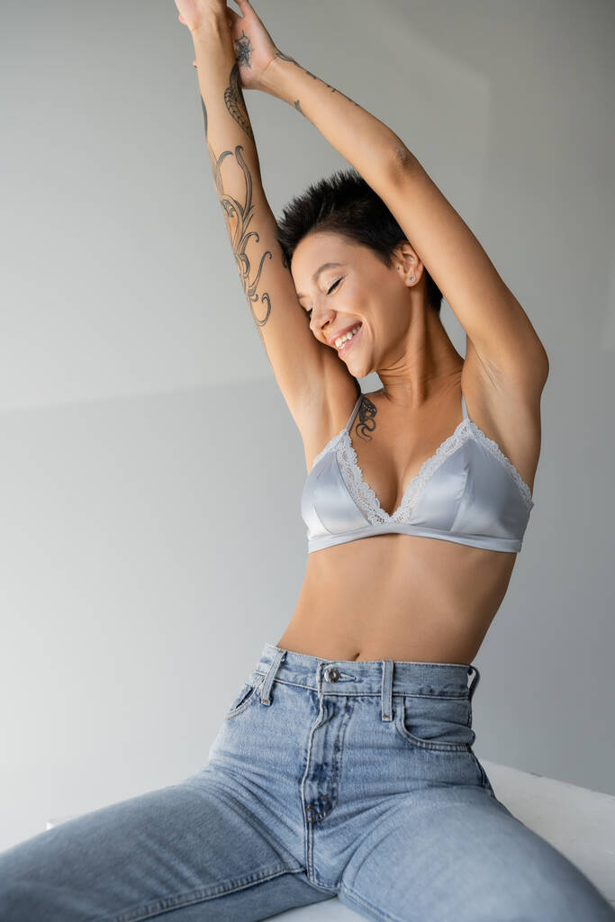 joyful tattooed woman in jeans and silk bra sitting on cube with raised hands and closed eyes on grey background - Foto, Bild