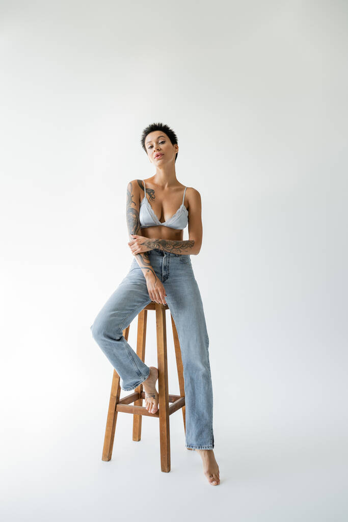 πλήρες μήκος της σαγηνευτικής ξυπόλητη γυναίκα σε τζιν και σουτιέν κάθεται σε ψηλό ξύλινο σκαμνί σε γκρι φόντο - Φωτογραφία, εικόνα