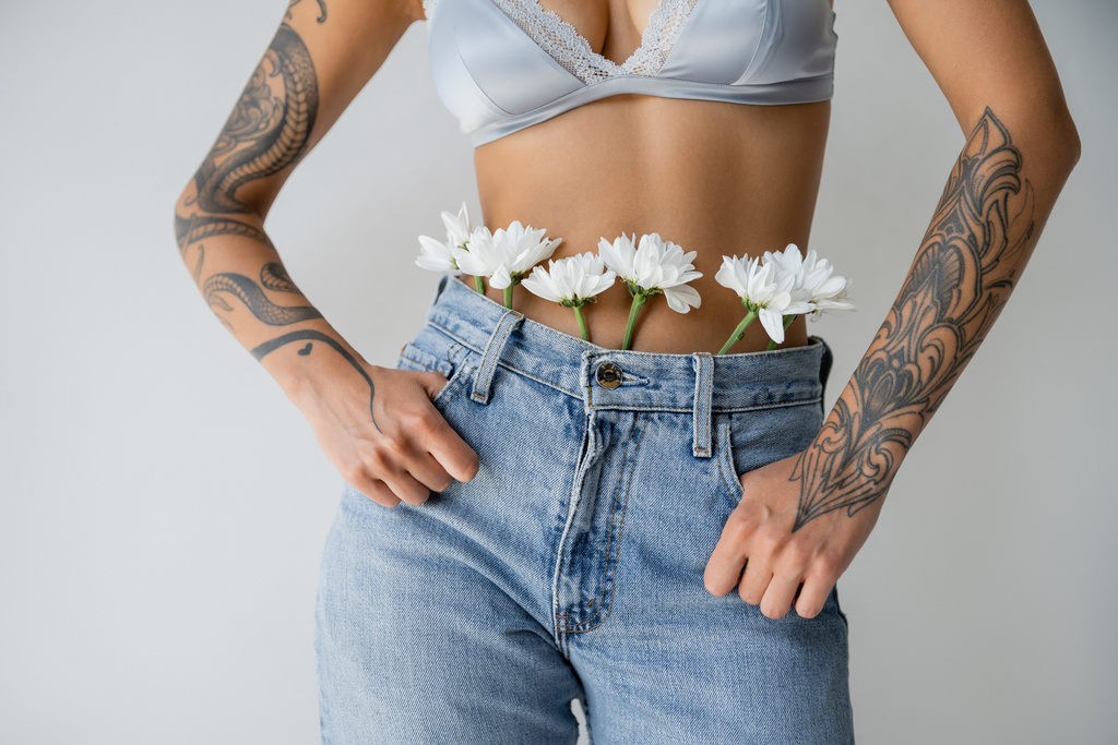 ořezaný pohled na tetovanou ženu v hedvábné podprsence a džínách s bílými květy stojící s palci v kapsách izolovaných na šedé - Fotografie, Obrázek