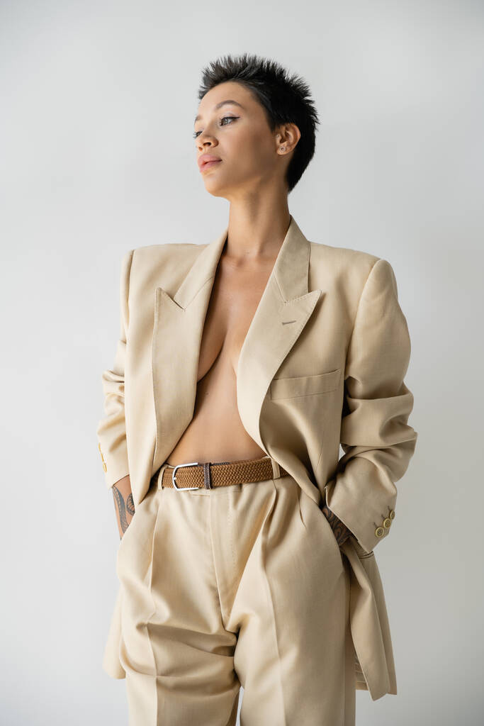 seductive woman in oversize blazer on shirtless body standing with hands in pockets and looking away isolated on grey - Photo, Image