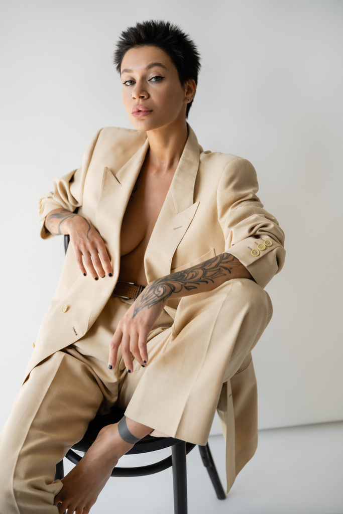 seductive tattooed woman in fashionable suit looking at camera while posing on chair on grey background - Foto, Bild