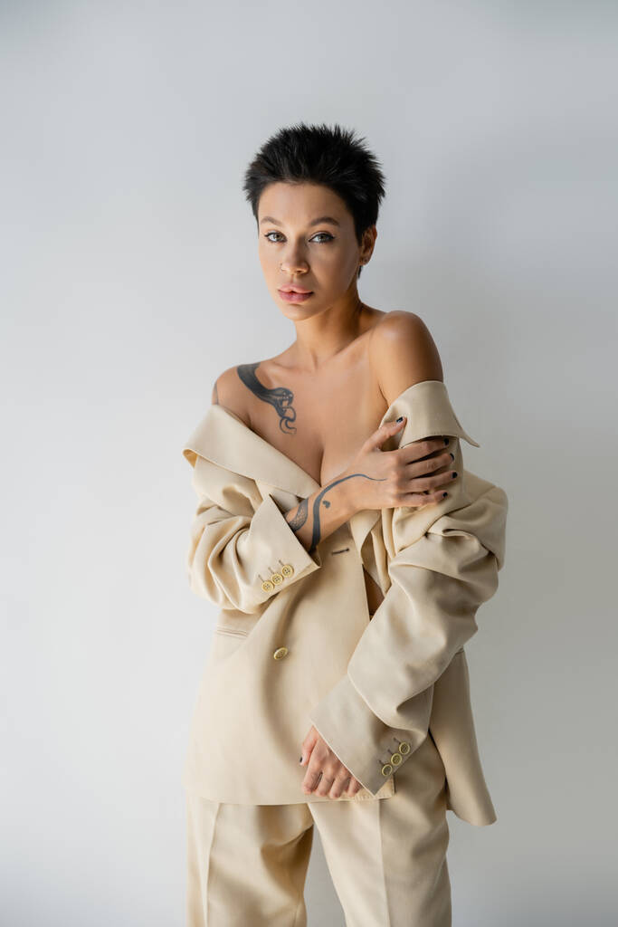 mujer tatuada con hombros desnudos posando en traje de gran tamaño mientras mira a la cámara aislada en gris - Foto, imagen