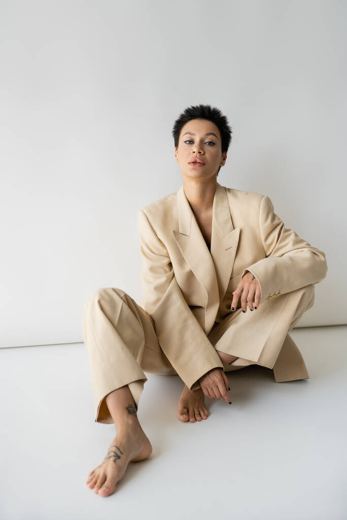 barefoot woman with short brunette hair looking at camera while sitting in beige suit on grey background - Photo, image