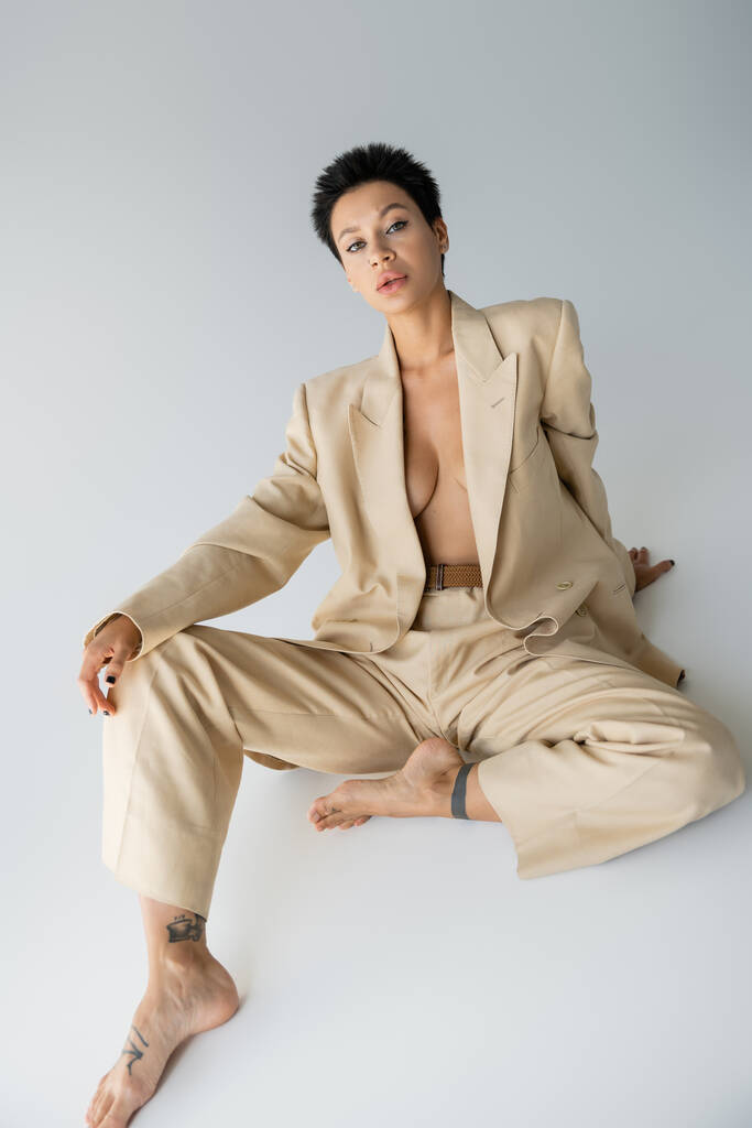 shirtless and barefoot woman in elegant pantsuit looking at camera while sitting on grey background - Foto, imagen