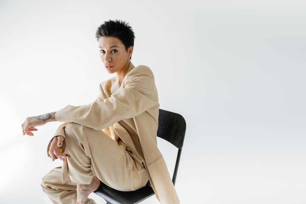 stylish brunette woman in beige suit looking at camera while sitting on chair on grey background - Foto, immagini