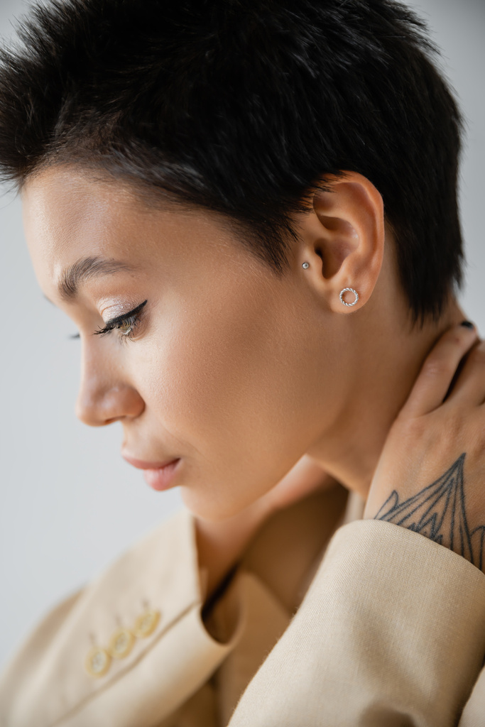 profile of young brunette woman with makeup and earrings posing with hands behind neck isolated on grey - 写真・画像