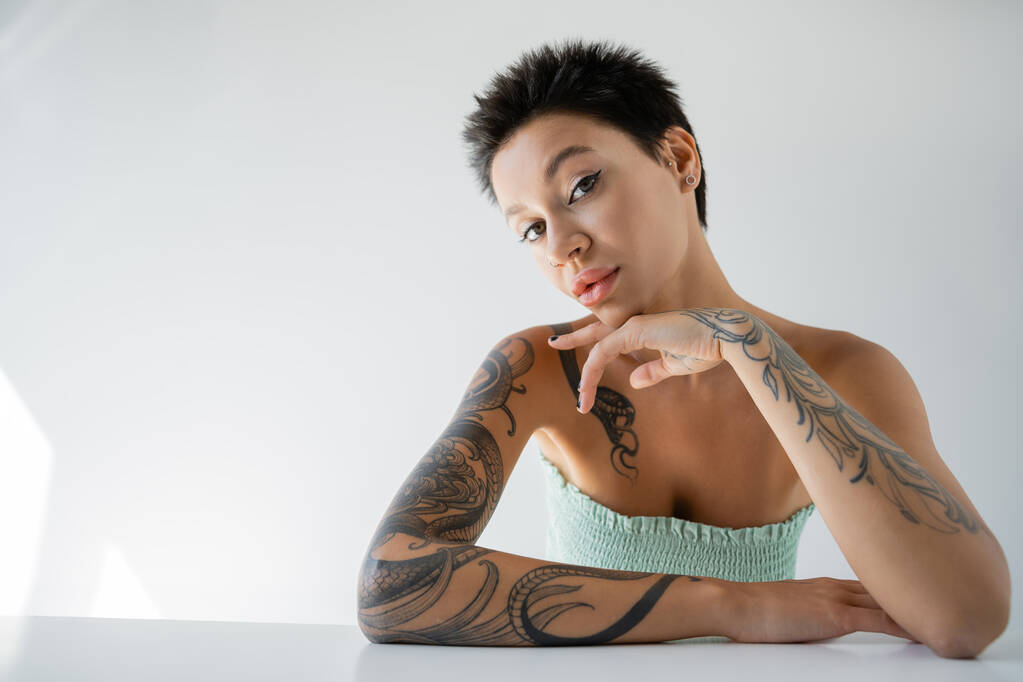 sensuale donna tatuata in top senza spalline seduta a tavola e guardando la fotocamera su sfondo grigio - Foto, immagini