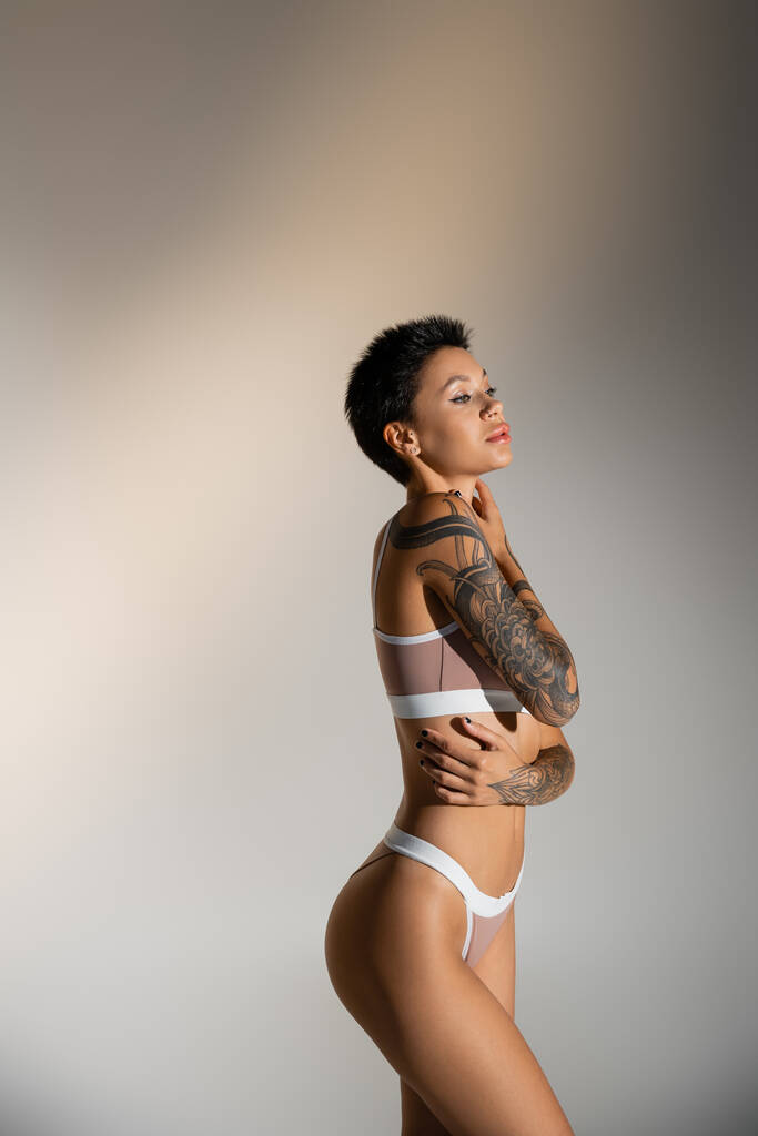 πλαϊνή όψη σέξι γυναίκα με τατουάζ στο εσώρουχο αγγίζοντας τέλειο σώμα σε γκρι φόντο - Φωτογραφία, εικόνα